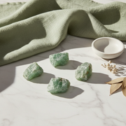 Four rough green crystal stones arranged on a white marble surface with a sage green linen cloth, a small white ceramic dish, and dried leaves as styling props.