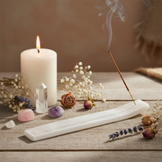 A white carved incense holder with a smoking incense stick, styled on a rustic wooden surface with a lit white pillar candle, crystals, dried flowers, and lavender sprigs.