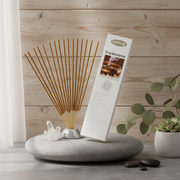 A white box of nitraj Frankincense incense sticks stands on a gray stone tray beside a fan of incense sticks in a silver holder, with a small plant and stones against a light wood plank background.