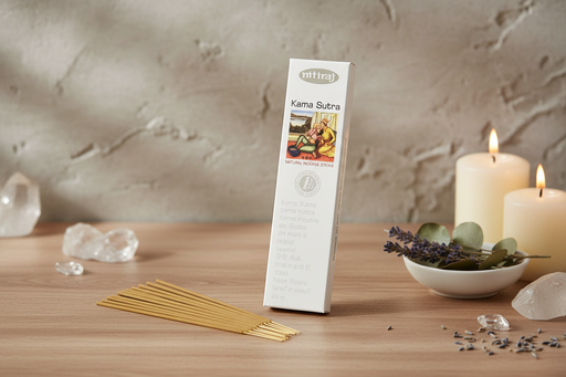 A boxed set of Kama Sutra incense sticks stands upright on a wooden surface, surrounded by lit white candles, crystals, dried lavender, and eucalyptus leaves against a textured beige wall.