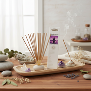 A boxed incense set labeled 'meditation' stands on a wooden tray beside several incense sticks, a smoking incense holder, crystals, dried flowers, and greenery in a softly styled indoor setting.