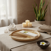 A styled tabletop scene with a round wooden tray holding a white rectangular box and several thin wooden sticks, surrounded by lit candles, crystals, a folded beige cloth, and a small bowl with a white flower. A potted snake plant sits in the background against a warm neutral wall.