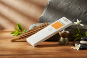 A boxed incense stick set labeled 'Sandalwood' by 'nitraj' lies on a wooden surface with loose incense sticks, decorative green leaves, white flowers, and a small metal incense burner against a stone backdrop.