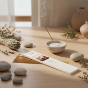 A boxed incense stick set sits on a light wood table beside a small bowl of incense powder, a smoking incense stick, stones, dried herbs, and ceramic vases in a softly lit interior setting.