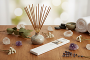 A reed diffuser with wooden reeds in a small speckled ceramic vase sits on a wooden table, alongside a white diffuser box, crystals, small white flowers, lavender sprigs, dark stones, a green bowl, and rolled towels in a softly blurred spa-like setting.