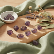 Ten polished ametrine  tumbled stones are arranged on a light wooden board, surrounded by a sage green fabric, a small white ceramic dish containing three stones, and dried lavender flowers.
