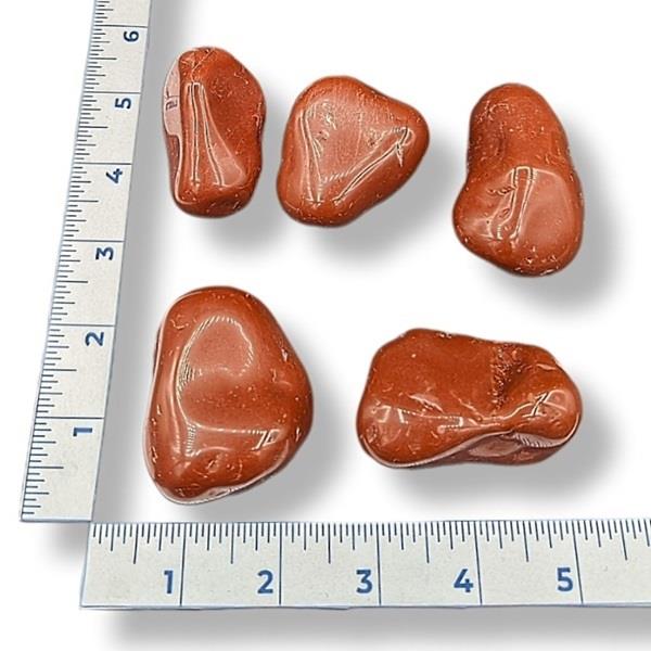 A collection of red tumbled jasper stones of various shapes, placed next to a ruler for size comparison.