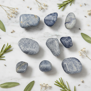 A set of polished blue and gray river stones arranged on a white marble surface with green leaves and small white flowers as decorative props.