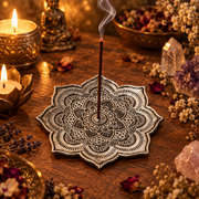 A single incense stick stands upright on a decorative mandala-shaped metal holder on a wooden table, surrounded by lit candles, dried flowers, crystals, and small decorative figurines in a warm, atmospheric setting.