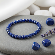 A beaded stretch bracelet made of polished dark blue lapis lazuli stones, displayed on a gray stone plate with loose lapis stones and a sprig of lavender nearby.