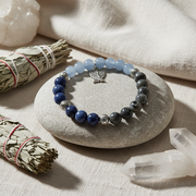 A beaded stretch bracelet with light blue, dark blue, and gray beads, accented by silver-toned spacer beads and a small butterfly charm, displayed on a smooth gray stone surrounded by natural props including a smudge bundle, crystals, and linen fabric.
