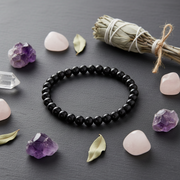 A black beaded bracelet arranged in a circle on a dark gray surface, surrounded by decorative stones, a dried sage bundle, and a clear crystal point.