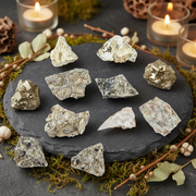 A set of natural mineral and crystal specimens arranged on a dark slate tray, surrounded by green moss, small branches with leaves, and lit candles in glass holders.