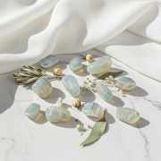 A set of polished light blue-green tumbled stones arranged on a white marble surface with draped white fabric and small dried flowers and leaves as styling props.
