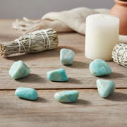 Seven polished turquoise-blue tumbled stones arranged on a rustic wooden surface, with a white pillar candle, dried sage bundle, and neutral fabric in the background.