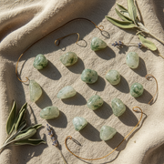A styled flat-lay of twelve polished green crystal tumbled stones arranged on a beige fabric background with decorative sage leaves and thin wire loops.