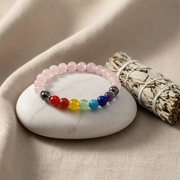 A beaded stretch bracelet with a rainbow of colored gemstone beads and dark metallic spacer beads, displayed on a smooth white stone next to a smudge bundle on a beige fabric background.