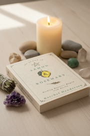 A book titled 'The Scent of Lemon Rosemary' by Raechel Henderson lies on a light wood surface, styled with a lit cream candle, smooth stones, a purple crystal cluster, and a sage bundle.