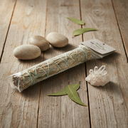 A packaged sage smudge bundle wrapped in clear plastic with a small paper tag, arranged on a rustic wooden surface with three smooth stones, green leaves, and a clear crystal cluster.