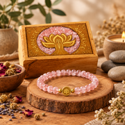 A pink beaded bracelet with a gold lotus charm is displayed on a wooden slice, with its engraved wooden presentation box behind it and spa-style props around it.