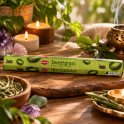 A box of HEM Sweetgrass incense sticks lies on a rustic wooden board surrounded by lit candles, green herbs, crystals, and plants in a warm, natural setting.