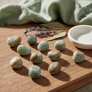 Ten polished aqua and beige jasper stones arranged on a wooden board, with a small white dish, dried lavender sprigs, and a sage green cloth in the background.