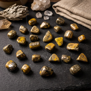 A collection of polished golden-yellow and brown tumbled stones arranged on a dark slate surface, with crystals and folded linen in the background.