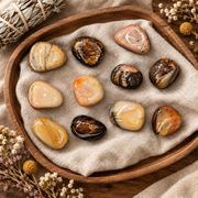 A set of polished tumbled gemstones in various earthy colors and patterns, arranged on a beige cloth inside a round wooden tray, with dried floral accents and a bundle of sage in the corners.