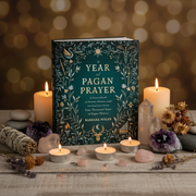 The book 'Year of Pagan Prayer' by Barbara Nolan is displayed on a rustic wooden surface surrounded by lit candles, crystals, a sage bundle, and dried flowers against a warm, glowing bokeh background.