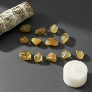 Ten small polished yellow tumbled stones arranged on a dark gray surface, with a white candle and a bundle of dried sage leaves as styled props.
