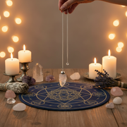 A hand holds a silver chain pendulum with a clear crystal quartz over a blue and gold circular crystal ball mat on a wooden table, surrounded by lit candles, crystals, and dried lavender.