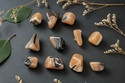 A set of polished tumbled gemstones with a peachy orange and cream body and black veining, arranged on a dark slate background with dried flowers and green leaves.