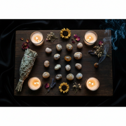 A styled flat-lay of a spiritual altar arrangement on a dark wooden board, featuring a white sage bundle, polished stones, dried flowers, a small acorn, and four lit tealight candles, with blue smoke rising on the right.