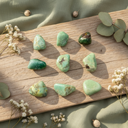 Nine polished green tumbled stones arranged on a rectangular wooden board, surrounded by small white flowers and green leaves on draped sage-green fabric.