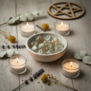 A styled tabletop scene with a white bowl filled with topaz crystals, surrounded by lit tealight candles, dried eucalyptus leaves, lavender sprigs, a brass incense burner with smoke, and a wooden pentagram symbol on a light wood surface.