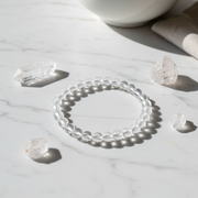 A clear beaded bracelet made of transparent round beads, arranged in a circle on a white marble surface with a few rough crystal stones nearby and soft shadows.
