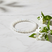 A beaded stretch bracelet made of white iridescent beads, styled on a white marble surface next to white flowers with green leaves.