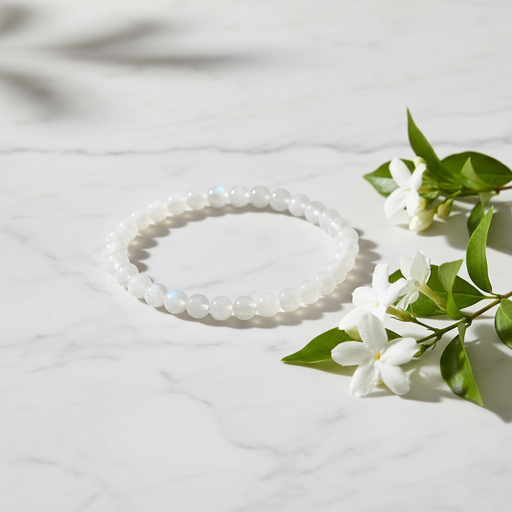 A beaded stretch bracelet made of white iridescent beads, styled on a white marble surface next to white flowers with green leaves.