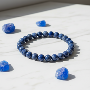 A beaded bracelet made of polished dark blue selenite beads, arranged in a circle on a white marble surface, surrounded by several rough blue crystal pieces.