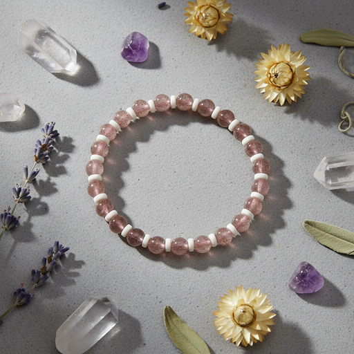 A beaded stretch bracelet made of pink translucent crystals and white spacer beads, arranged in a circle on a gray surface with crystals, dried flowers, and lavender sprigs around it.