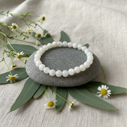 A white beaded bracelet made of smooth, polished round beads, displayed on a smooth gray stone. The stone is styled on a textured beige linen fabric, surrounded by green leaves and small white daisies.