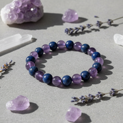 A beaded stretch bracelet made of alternating deep blue lapis lazuli and light purple amethyst round beads, styled on a gray surface with lavender sprigs, crystals, and a larger amethyst cluster.