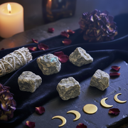 Five raw labradorites arranged on a black cloth with a white sage bundle, purple flowers, scattered rose petals, and gold crescent moon and full moon charms, with a lit candle in the background.