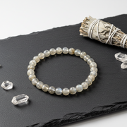 A beaded stretch bracelet made of polished grayish-white labradorite beads, displayed on a black slate tray with crystal points and a white sage bundle.