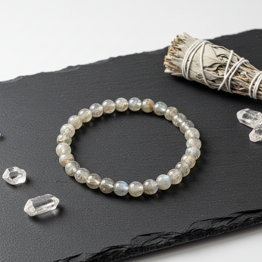 A beaded stretch bracelet made of polished grayish-white labradorite beads, displayed on a black slate tray with crystal points and a white sage bundle.