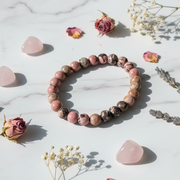 A beaded stretch bracelet made of polished pink and brown stone beads with some dark marbled sections, arranged in a circle on a white marble surface with dried roses, crystals, and small white flowers around it.
