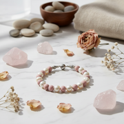 A beaded bracelet with pink and white beads and a silver-toned clasp, styled on a white marble surface with pink stones, dried flowers, pebbles, and a folded beige fabric in the background.