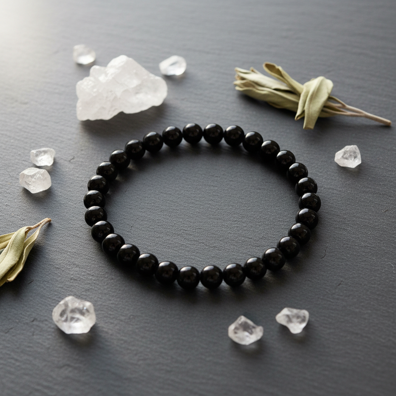 A black beaded bracelet arranged in a circle on a dark gray slate surface, surrounded by clear crystal clusters and dried green leaves.