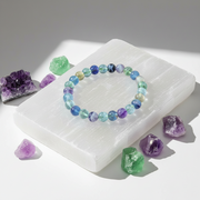 A beaded stretch bracelet made of round, translucent gemstone beads in shades of blue, purple, green, and clear, displayed on a white rectangular slab with purple and green crystals around it.