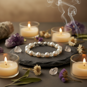 A beaded bracelet made of white pearl-and labradorite beads, displayed on a dark slate tray surrounded by lit candles, crystals, dried flowers, and leaves.